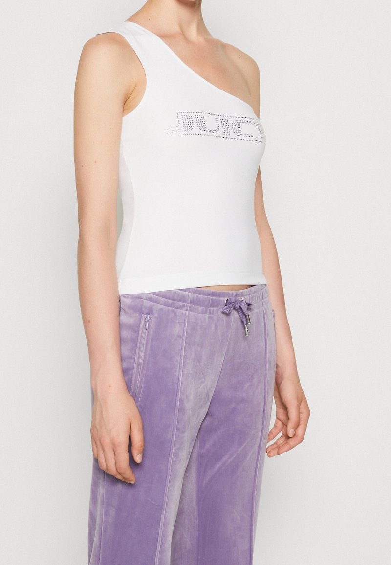 Bílý top na jedno rameno s přiléhavým střihem a grafickým potiskem "JUICE" vyloženým z kamínků, doplněný o světle fialové velurové kalhoty se zipovými kapsami.