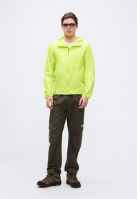 Νεαρός άνδρας που στέκεται φορώντας φωσφοριζέ κίτρινο μπουφάν, σκούρο παντελόνι πεζοπορίας, μπότες πεζοπορίας και σκουρόχρωμα γυαλιά ηλίου σε λευκό φόντο.