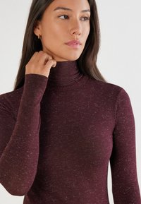 Weinroter Rollkragenpullover mit langen Ärmeln, aus einem weichen, strukturierten Stoff mit dezenten Sprenkeln gefertigt. Eng anliegendes Design am Hals.