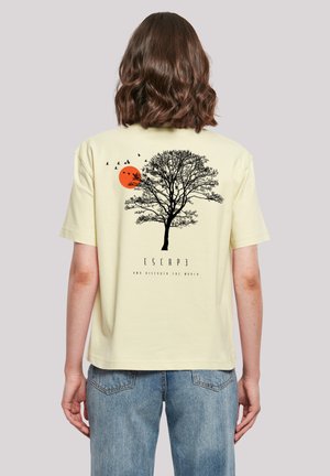 Person with shoulder-length brown hair wearing light yellow T-shirt with black tree, orange sun, flying birds, and text "ESCAPE" on back.