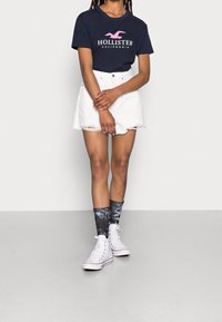 Marinblå t-shirt med rosa "Hollister"-logga, vita denimshorts, svart och vit batikstrumpor samt vita hög-top sneakers.