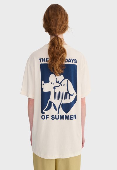Personne aux longs cheveux roux attachés en queue de cheval, portant un t-shirt crème avec un motif bleu de deux chiens et le texte "LES JOURS D'ÉTÉ" au dos.