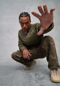 Chaqueta bomber verde oliva con cremallera, pantalones de camuflaje marrón con patrón y zapatillas color beige. El modelo posa en una posición agachada con la mano levantada.