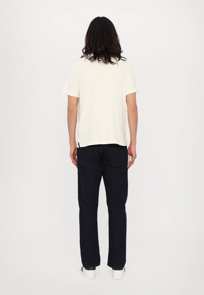 Witte T-shirt met korte mouwen, gecombineerd met donkerblauwe jeans, gezien van achteren. T-shirt heeft een rechte snit; de jeans hebben een klassieke pasvorm.