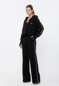 Czarna bluza z kapturem na zamek z logo, długie rękawy i kieszenie z przodu w zestawie z dopasowanymi spodniami o szerokich nogawkach z bocznymi akcentami logo. Miękka tekstura materiału.