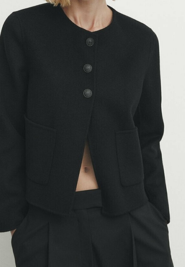 Femme portant un cardigan noir boutonné avec deux poches avant et un pantalon noir assorti, laissant apparaître une petite section du ventre.