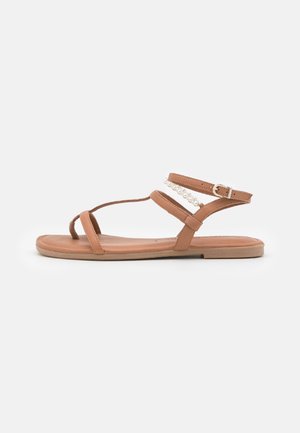 Dames flad tan sandal med tynde stropper, ankelspænde og dekorativ perledetalje på en stropp, vist mod hvid baggrund.