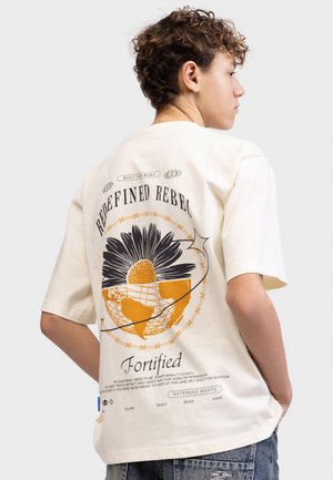 Młoda osoba z krótkimi kręconymi włosami, ubrana w kremowy t-shirt z dużą grafiką słonecznika i globu oraz tekstem na plecach.