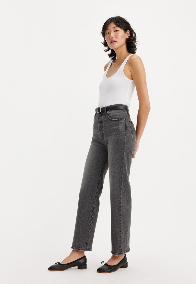 Jean noir taille haute avec une jambe droite, comprenant des passants de ceinture et des poches latérales. Porté avec un débardeur blanc et des ballerines noires.