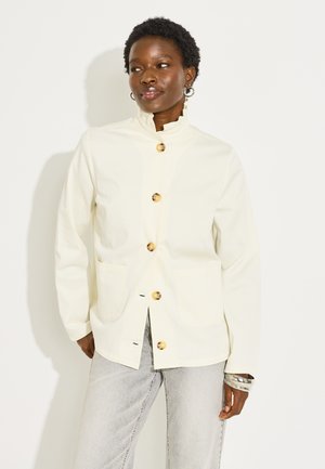 Mujer con una chaqueta blanca abotonada con grandes botones de madera y dos bolsillos delanteros, combinada con vaqueros gris claro y pulseras plateadas.