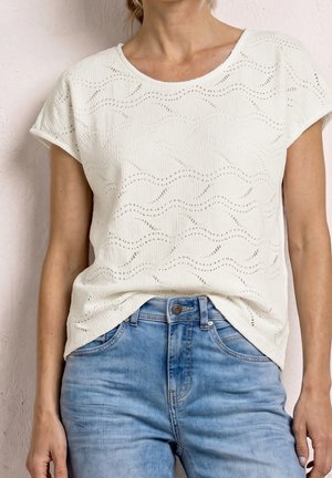 Frau trägt ein weißes, kurzärmeliges, strukturiertes Oberteil mit Wellenmustern, das in hellblaue hoch taillierte Jeans gesteckt ist.