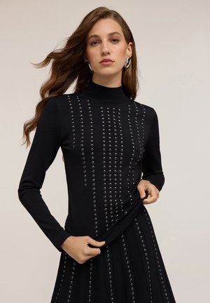 Maglione nero aderente con un colletto alto, caratterizzato da file verticali di borchie circolari per un effetto tattile e di contrasto. Tessuto a maglia morbida.