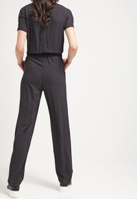 Musta haalari lyhyillä hihoilla, joustavalla vyötäröllä ja leveillä lahkeilla. Sileä pinta ja minimalistinen tyyli. Rentoa jalkineita näkyvissä alhaalla.