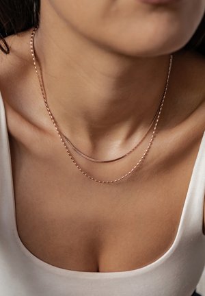 Doppellagige Halskette aus Roségold mit einer glatten Kette und einer strukturierten Kette, die auf nackter Haut über einem hellen Oberteil liegt.