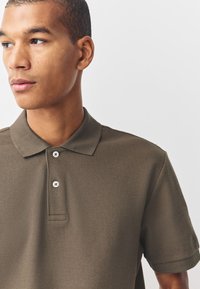 Joven con cabello corto que lleva una camisa polo texturada de color verde oliva, mirando a la izquierda contra un fondo blanco sencillo.