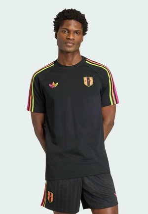 Homme portant une tenue de sport noire avec des rayures roses et jaunes, logos Adidas et FPF, debout les mains derrière le dos sur un fond clair.