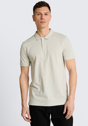 Młody mężczyzna w jasnobeżowej koszulce polo z krótkim rękawem i czarnych spodniach, stojący na jednolitym jasnoszarym tle.