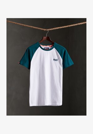 Maglietta bianca con maniche raglan verde scuro appesa a una gruccia di legno da una corda marrone spessa su uno sfondo scuro.