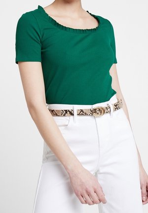 Camiseta de canalé verde con escote en forma de ondas, combinada con pantalones blancos y un cinturón texturizado con estampado de serpiente que tiene una hebilla redonda dorada.