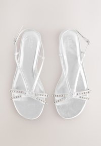Sandales métalliques argentées à semelle plate, avec des brides croisées ornées de strass pour embellissement.