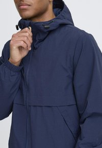 Navy blue lightweight jacket with a hood, elastic cuffs, and a zippered front. Features a horizontal seam and hand pockets. Smooth texture.