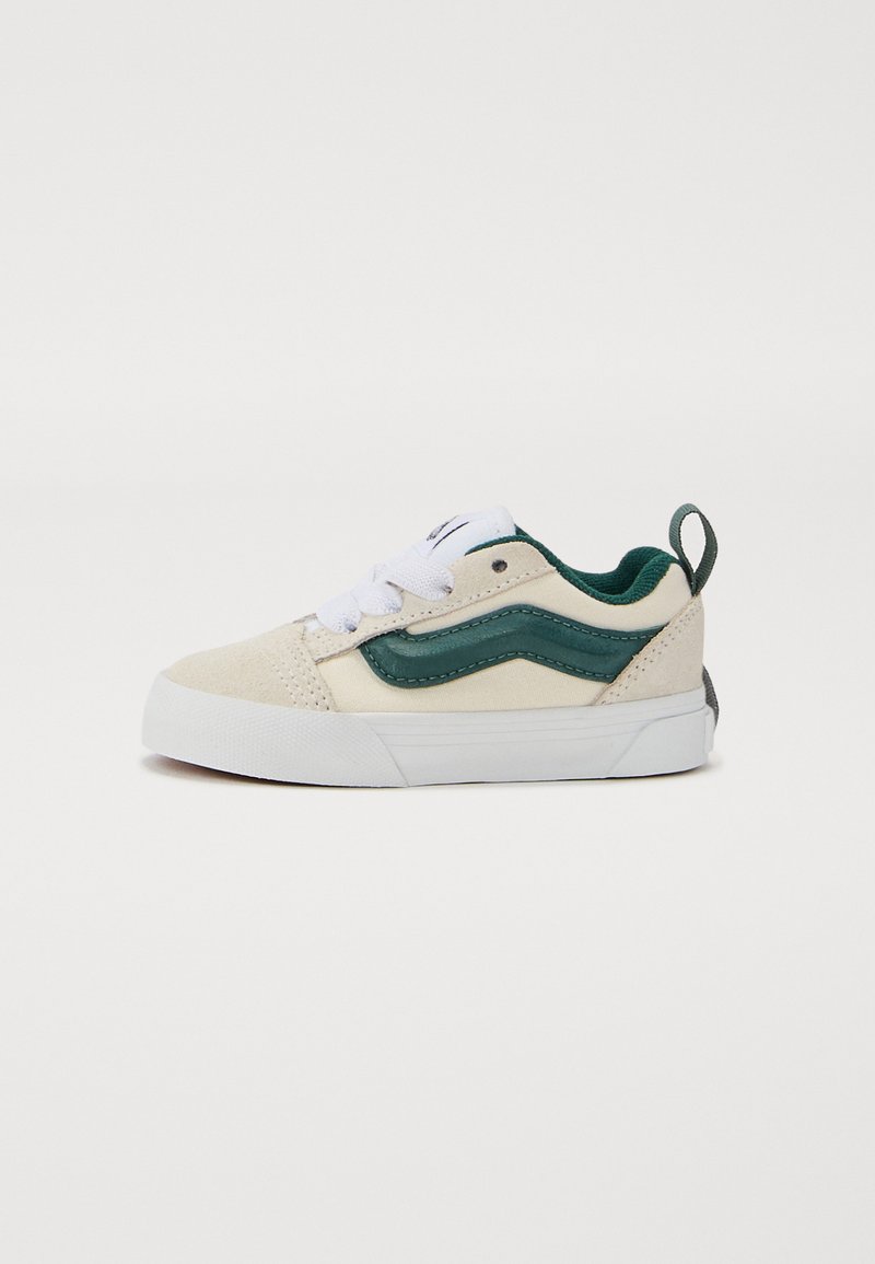 Tan canvas sneaker with green leather accents, white laces, and rubber sole. Features a side logo and a small heel tab for easy wear.