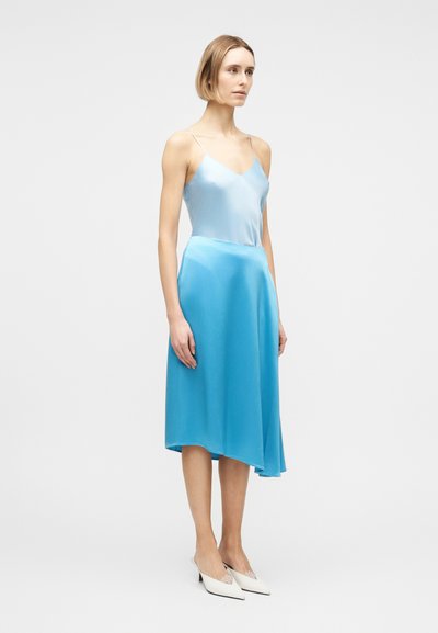 Γυναίκα που στέκεται, φοράει ανοιχτό μπλε σατέν τοπ καμισόλ και αντίστοιχο μίντι φούστα, με λευκά παπούτσια με μυτερή μύτη, σε απλό φόντο.