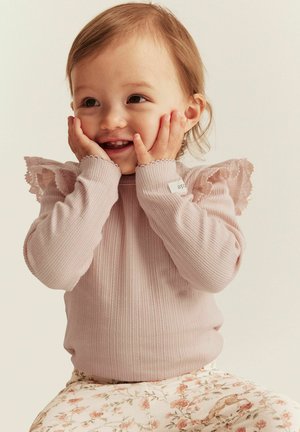 Lyserød ribbet skjorte med lange ærmer og blondekant detaljer på skuldrene, parret med bukser med blomster-mønster i lyse farver og illustrationer.