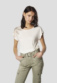 Balta īsroku T-krekls ar lapu rakstu uz kreisā pleca, kopā ar olīvu zaļām cargo biksēm ar daudzām kabatām un jostu.