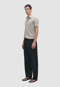 Polo beige, maniche corte, di colore uniforme; pantaloni larghi neri; infradito. Il modello posa in posizione eretta su uno sfondo grigio chiaro.