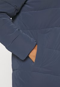 Marineblå pufferjakke med quiltet design, isolerende stof og en sidelomme. Mansjetten har en let forhøjet tekstur for ekstra varme.