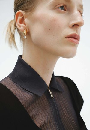 Pendiente en tono dorado con un diseño texturizado, que presenta un acento de gota clara. Modelo vistiendo una prenda negra con un detalle de cuello translúcido.