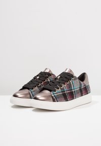 Zapatilla de cuadros con parte superior de tela negra, puntera metálica bronce y suela de goma blanca. Cuenta con cordones negros y ojales dorados.