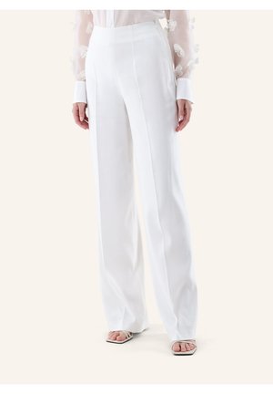 Donna che indossa pantaloni bianchi a vita alta e gamba larga, una camicetta trasparente con maniche con applicazioni floreali e sandali bianchi con punta aperta.
