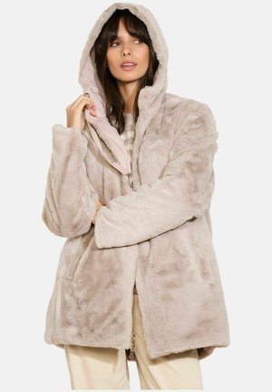 Cappotto con cappuccio in pelliccia sintetica di un beige chiaro, caratterizzato da una texture morbida, chiusura con zip e un rivestimento interno a contrasto. Abbinato a un maglione lavorato a maglia.