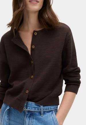 Frau trägt dunkelbraune, geknöpfte Strickjacke und blaue hoch taillierte Jeans, Hand in der Tasche, vor weißem Hintergrund.