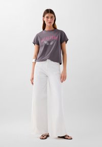 T-shirt en coton gris avec le texte "SCALPERS" en rose et un motif d'aile. Pantalons en denim blancs larges. Sandales plates noires avec des éléments décoratifs.