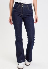 Ciemnoniebieskie dżinsy z rozszerzanymi nogawkami mają wysoki stan z dwoma guzikami, kontrastowe szwy oraz kieszenie z przodu, zestawione z białymi trampkami.