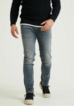 Uomo che cammina indossando jeans blu sbiaditi, maglione nero a maglia e sneaker bianche e nere su uno sfondo neutro.