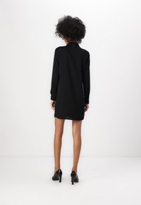 Robe noire à manches longues avec un col, arrivant mi-cuisse, faite d'un tissu lisse. Présente des accents de boutons sur les poignets et un design minimaliste.