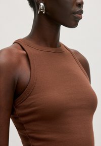 Bruine geribbelde mouwloze top met een ronde halslijn en een strak ontwerp. Zilveren ringoorbel zichtbaar aan het oor van het model. Gladde stoftextuur.