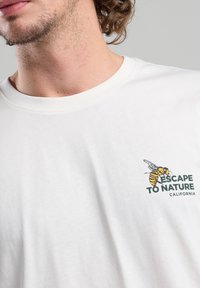 Valkoinen puuvillateeppari, jossa on grafiikka, jossa on mehiläinen ja teksti "PAETAA LUONTOON KALIFORNIA" vihreällä. Lyhyet hihat, pyöreä kaula-aukko.