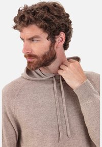 Sweat à capuche en maille beige avec cordon de serrage, manches raglan et poignets côtelés. Le tissu semble doux avec une texture lisse, offrant une coupe décontractée.