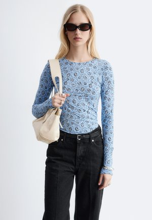 Kvinde med langt blondt hår iført sorte solbriller, en blå blomstret langærmet top, sorte bukser og bærer en beige skuldertaske.