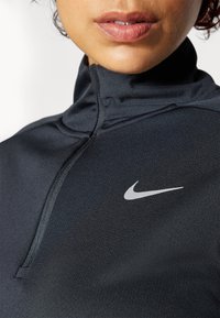 Czarna sportowa bluzka na zamek z wysokim kołnierzem, z srebrnym logo Nike na piersi. Materiał jest gładki i dopasowany, podkreślający smukły design.