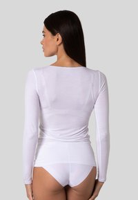 Donna con lunghi capelli scuri, che indossa un top bianco attillato a maniche lunghe e slip bianchi, vista di spalle su uno sfondo semplice.