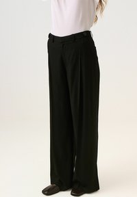 Fekete, bő lábszárú nadrág, redős elejével, gombos záródással, sima textúrával, fehér felsővel és sötét cipővel viselve.