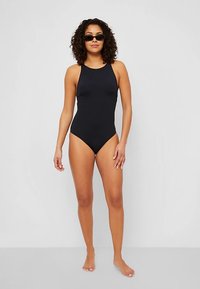 Μαύρο ολόσωμο μαγιό με ψηλό λαιμό, που διαθέτει εφαρμοστή σιλουέτα και λεία υφή. Το μοντέλο φορά σκοτεινά γυαλιά ηλίου και στέκεται ξυπόλητο.