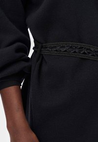 Czarny materiałowy element garderoby o fakturowanej powierzchni, z detalem w postaci czarnego, tkackiego pasa w talii i elastycznymi mankietami.