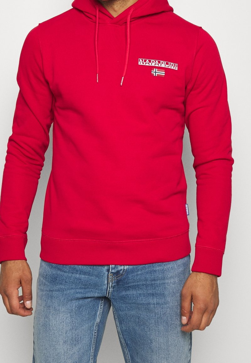 Homme portant un sweat à capuche rouge vif avec des cordons blancs et un petit logo Napapijri ainsi qu'un patch drapeau sur la poitrine, associé à un jean bleu.